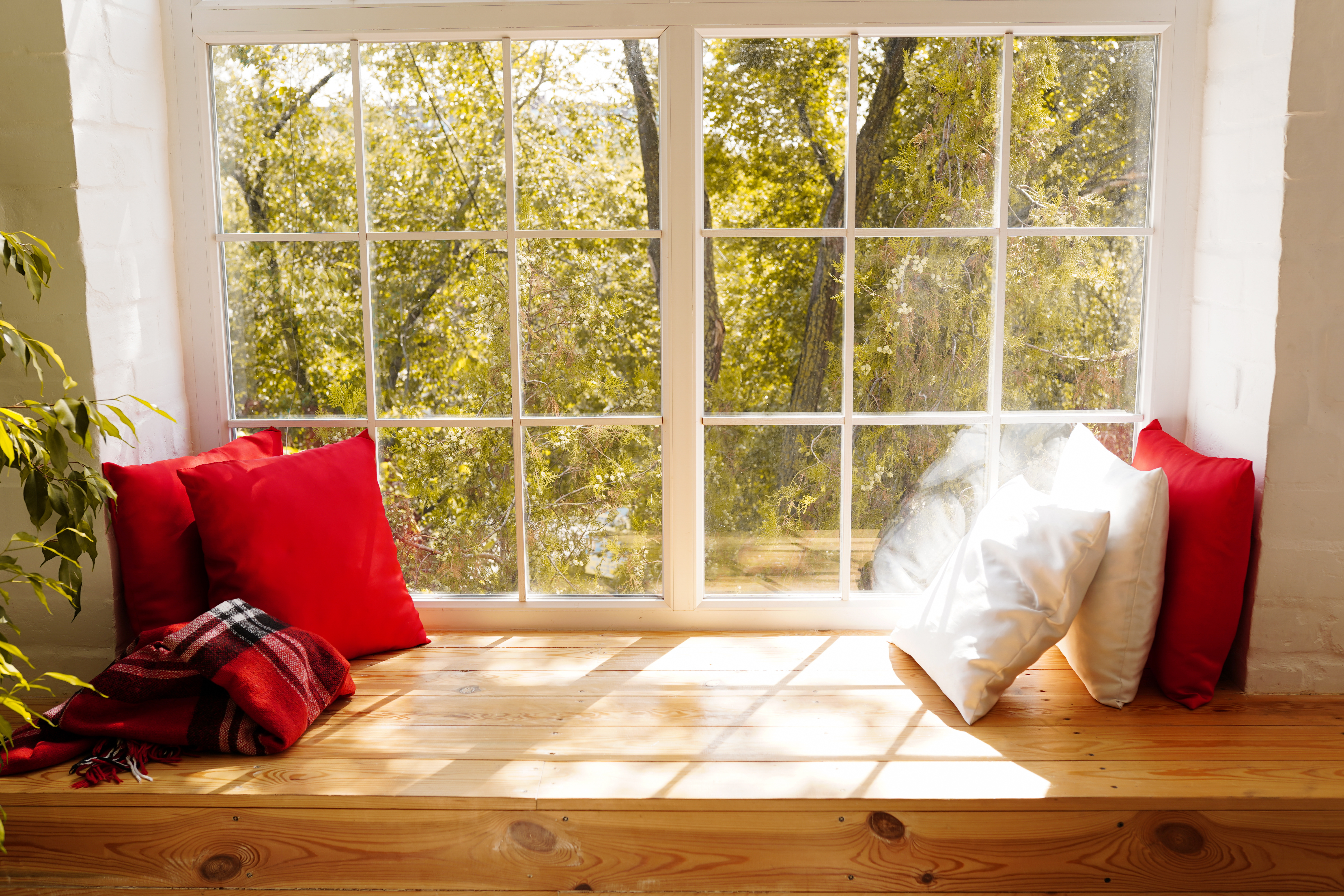 Alféizar ancho de madera maciza como asiento con cojines rojos y blancos ante ventana con travesaños, vista al jardín