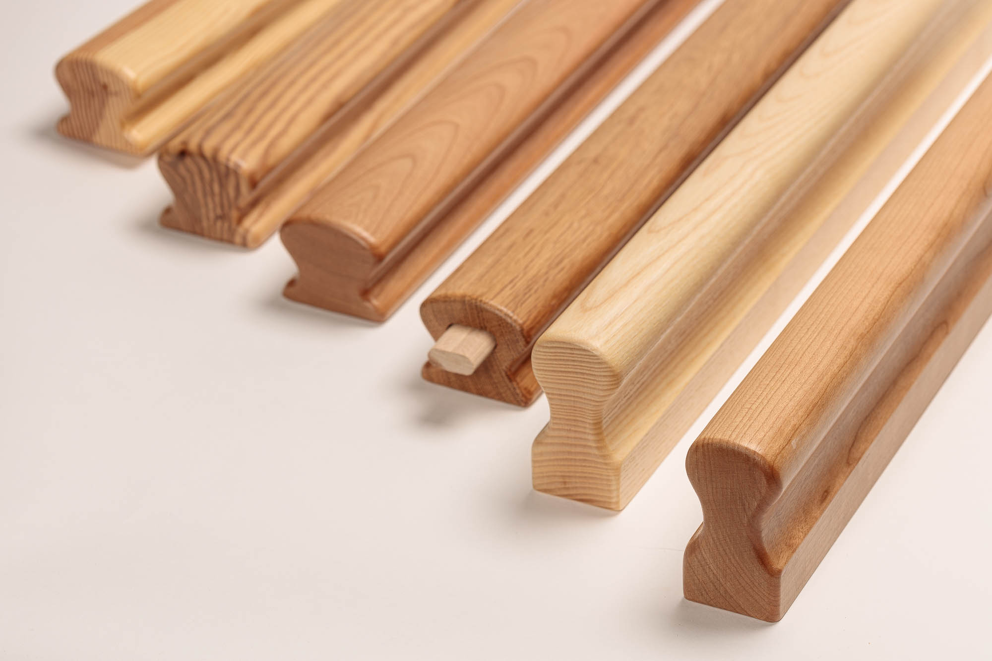 Pasamanos Omega de madera de seis especies uno junto a otro, maderas claras a rojizas, perfil Omega con hueco de agarre