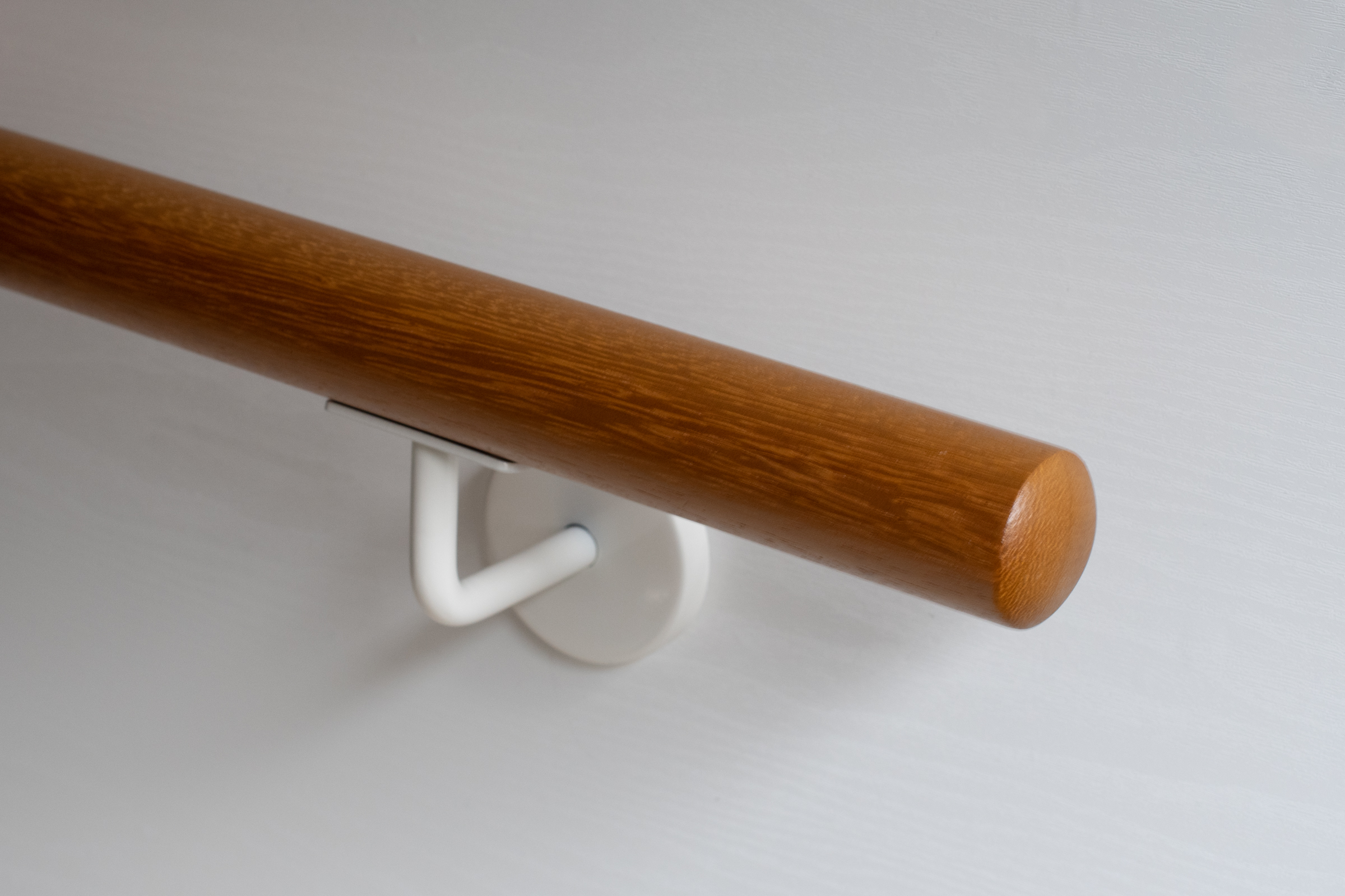 Pasamanos redondo de iroko con soporte lacado en blanco en pared blanca, madera marrón dorado, detalle del extremo