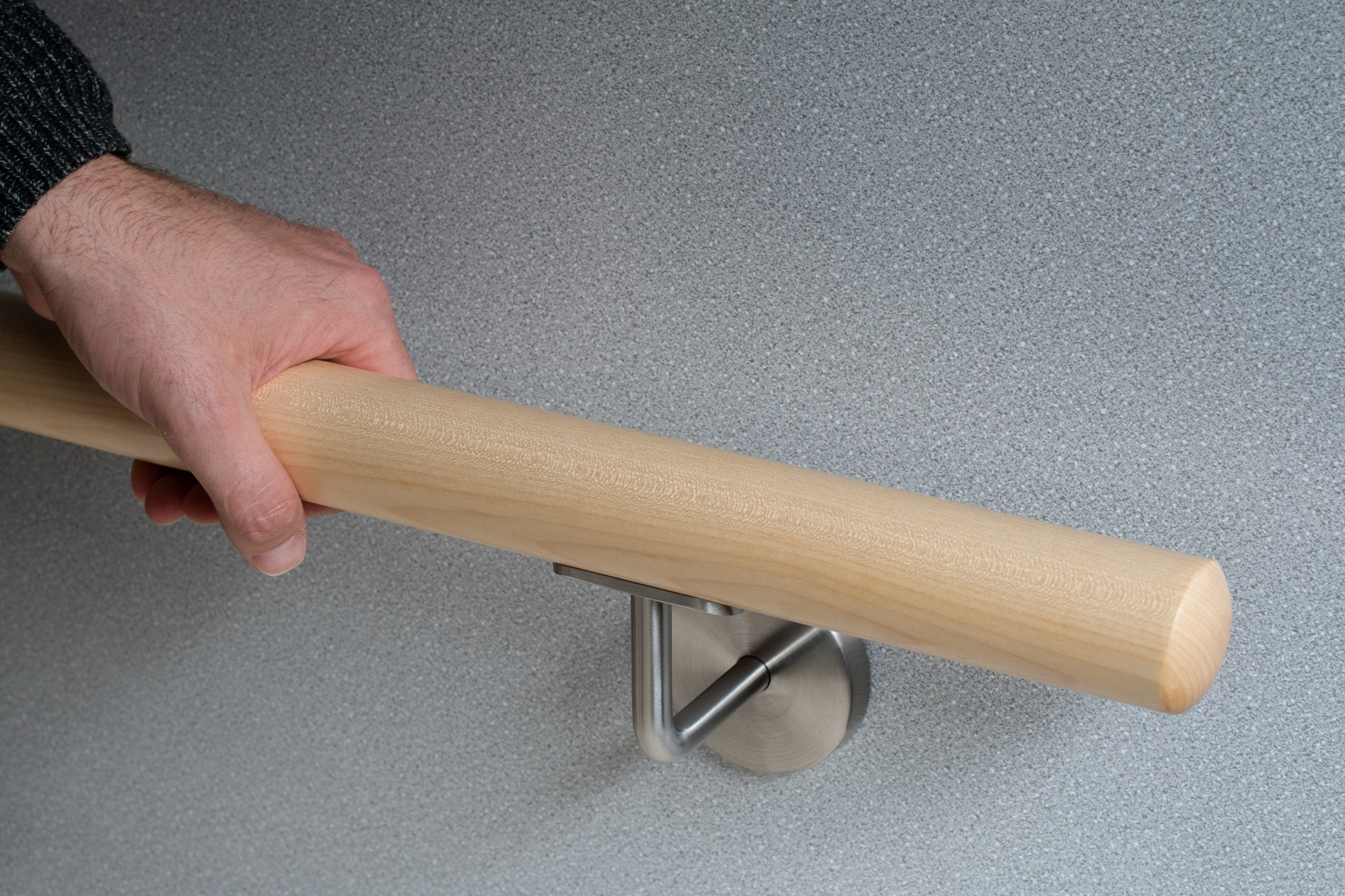 Pasamanos redondo de arce con soporte de acero inoxidable ante pared gris, una mano agarra el pasamanos claro, primer plano