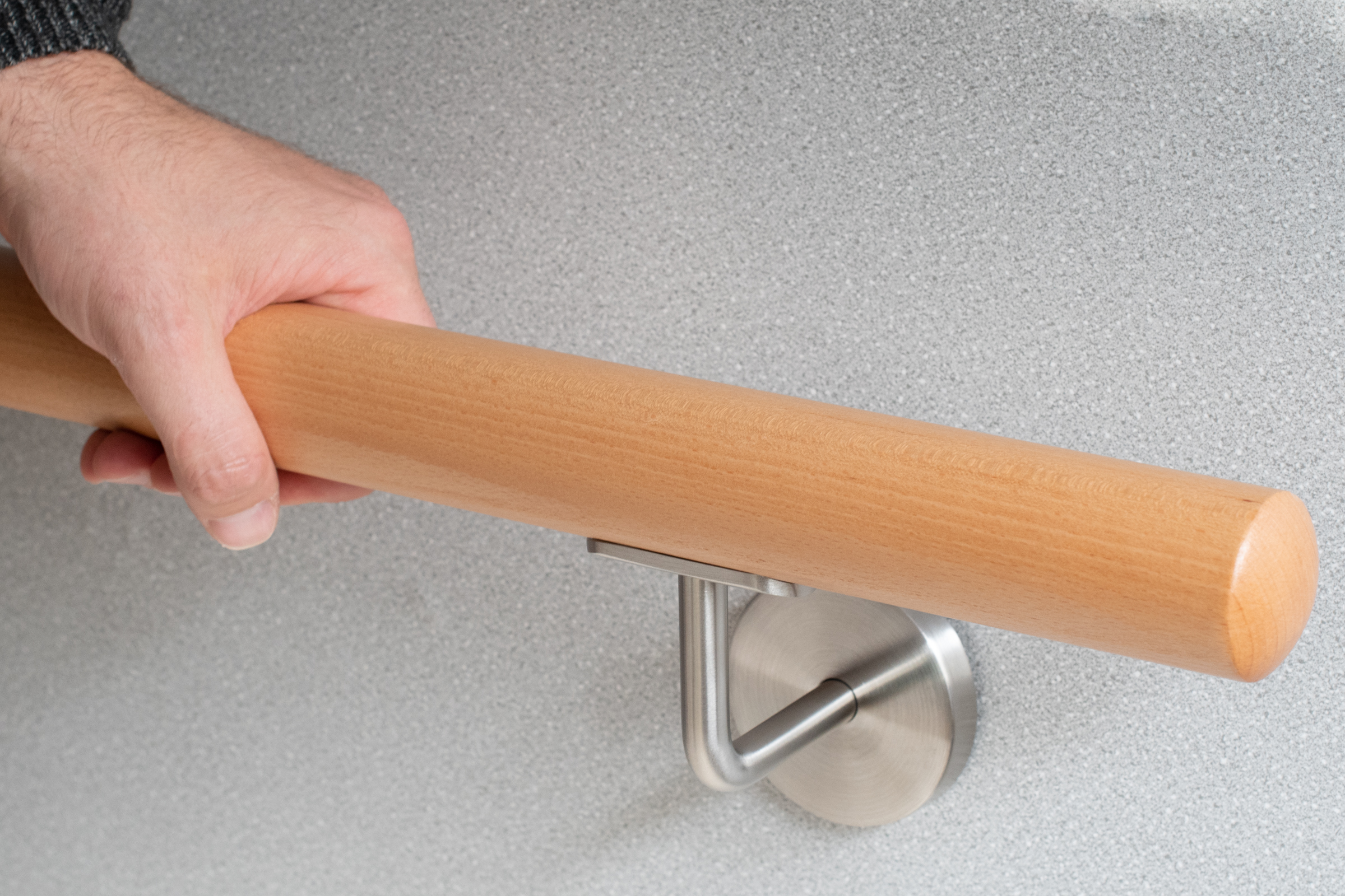 Pasamanos redondo de haya en pared gris con soporte de acero inoxidable, una mano agarra el pasamanos, detalle del agarre