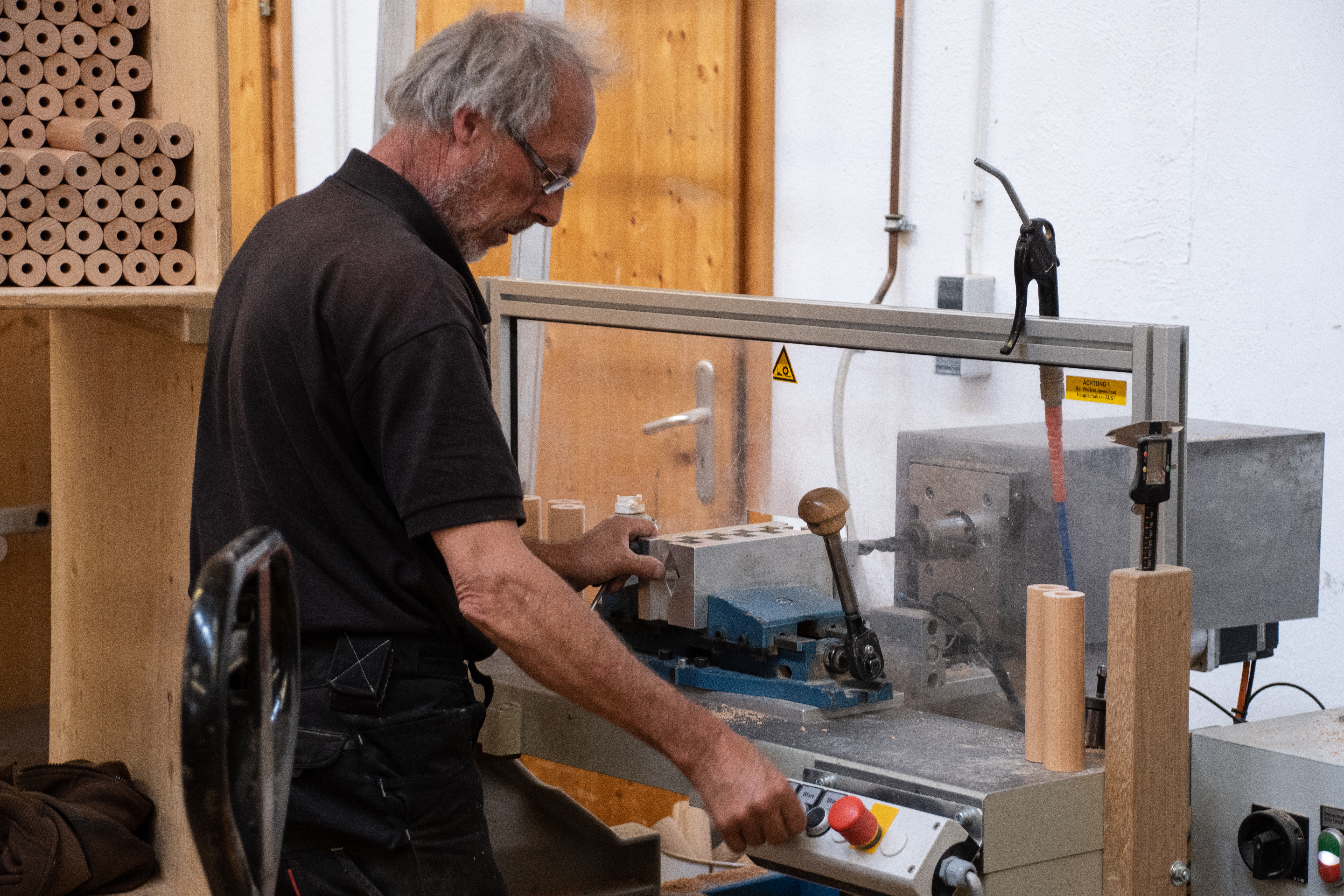 Fabricación de pasamanos: empleado taladra una pieza de madera en el taladro del taller, barras redondas al fondo