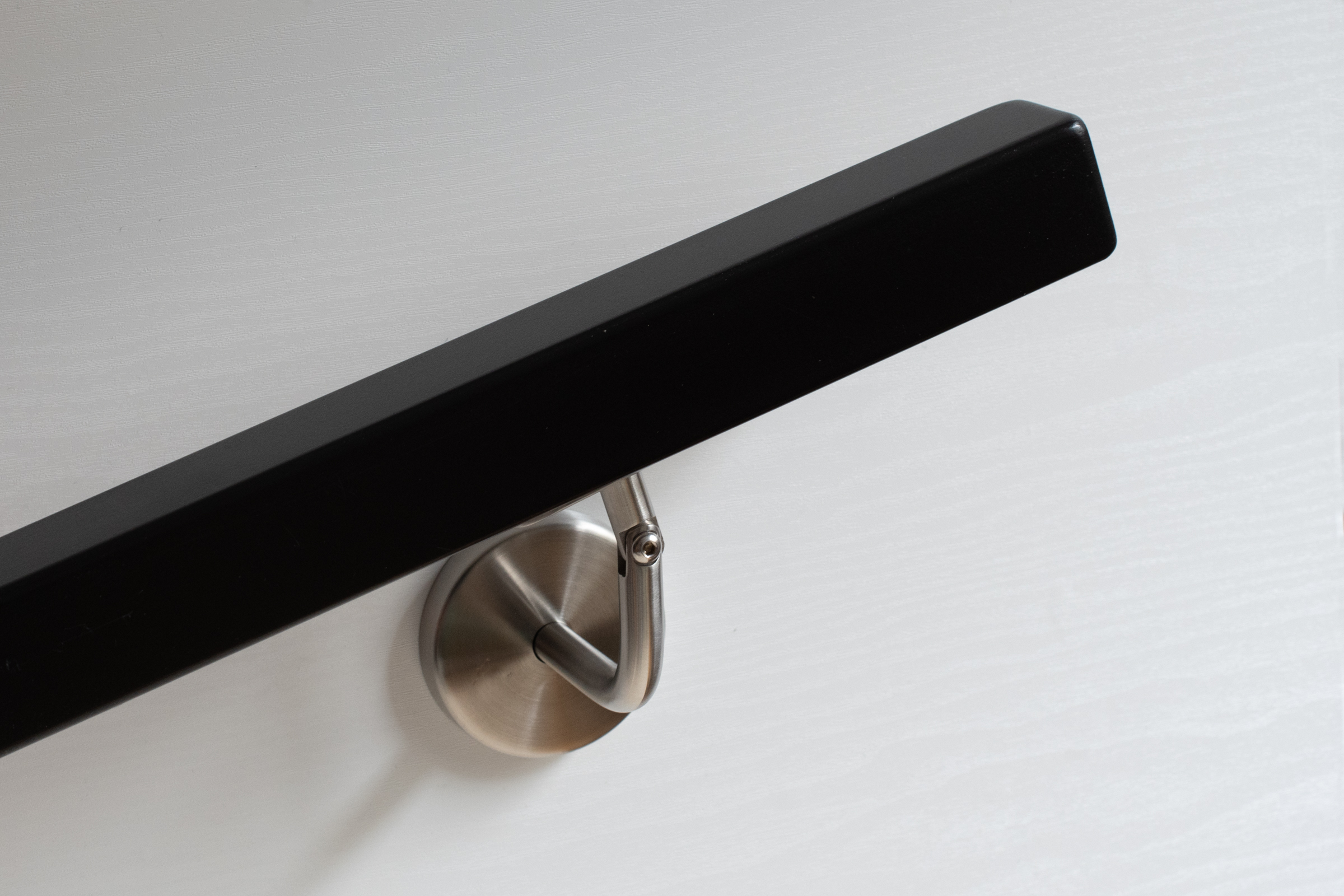 Pasamanos cuadrado lacado negro con soporte de acero inoxidable en pared de madera blanca, superficie mate, vista oblicua