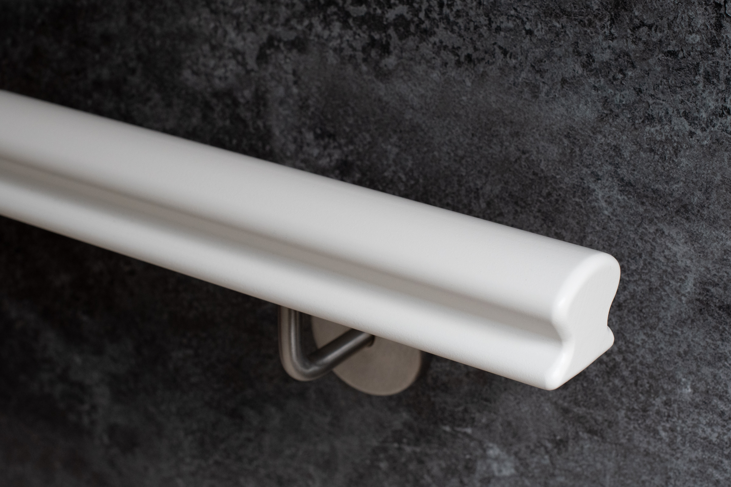 Pasamanos Omega de madera lacado en blanco con soporte de acero inoxidable ante pared gris oscuro, detalle del perfil