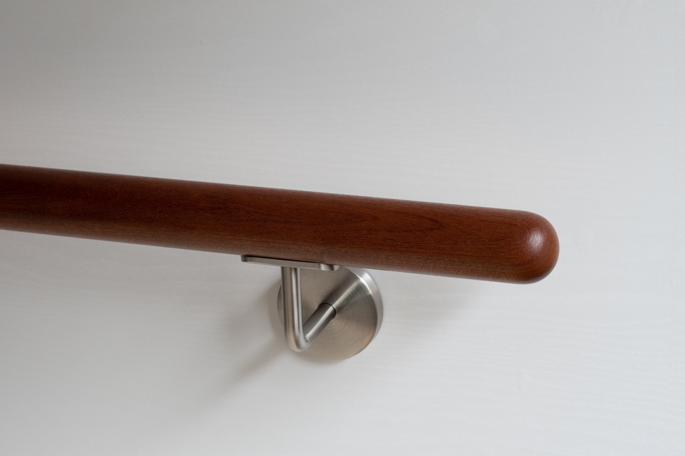 Pasamanos redondo de caoba con soporte de acero inoxidable en pared blanca, madera marrón rojiza, detalle del extremo