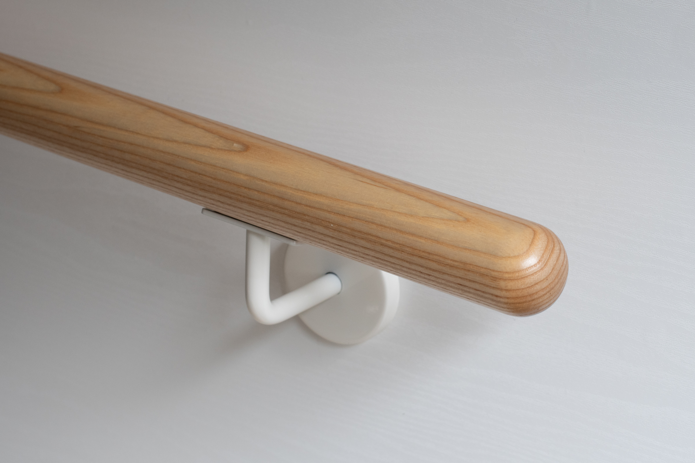 Pasamanos redondo de fresno con soporte lacado en blanco en pared blanca, madera clara de veteado marcado, primer plano