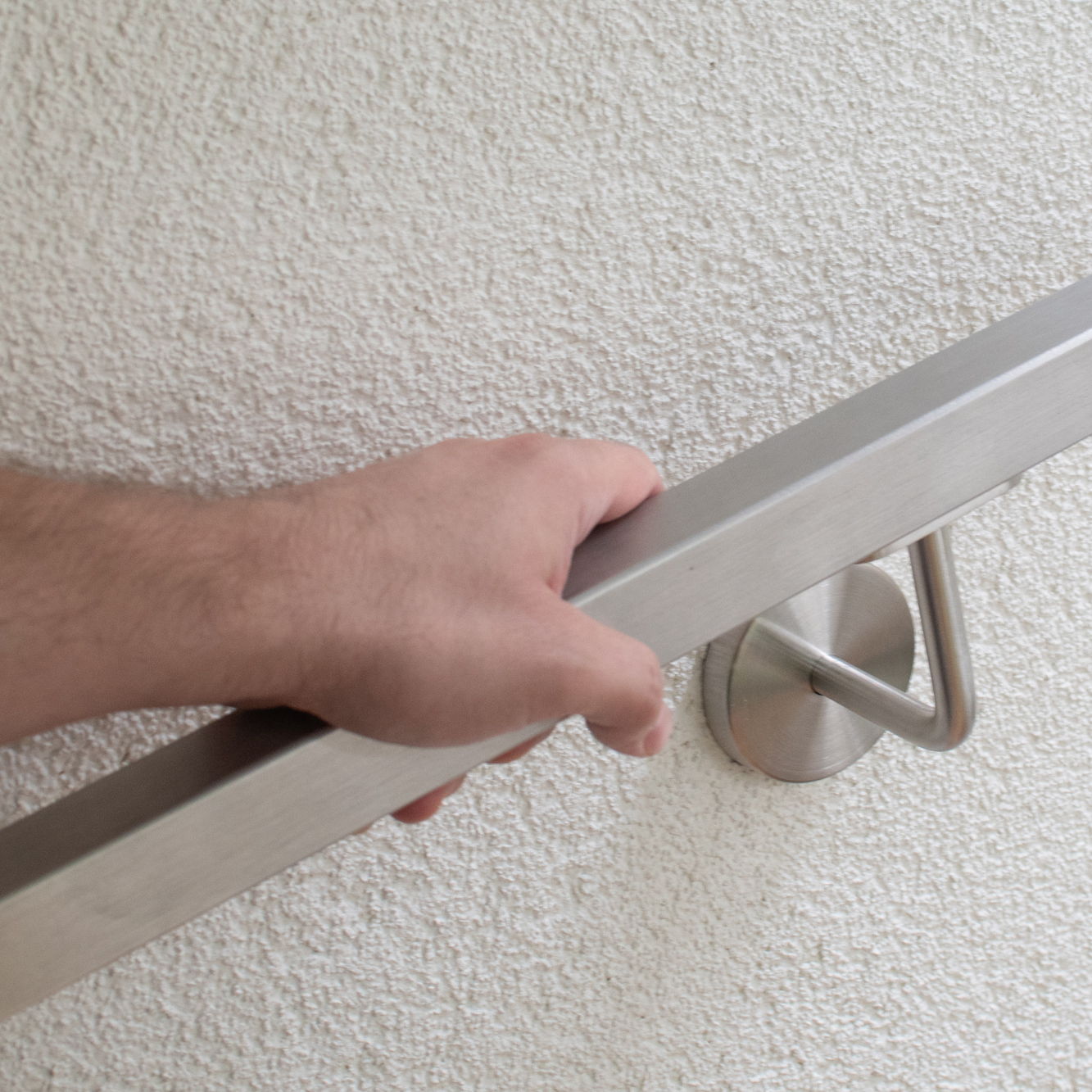 Pasamanos cuadrado de acero inoxidable en pared enlucida, una mano lo agarra, soporte de pared de acero con roseta