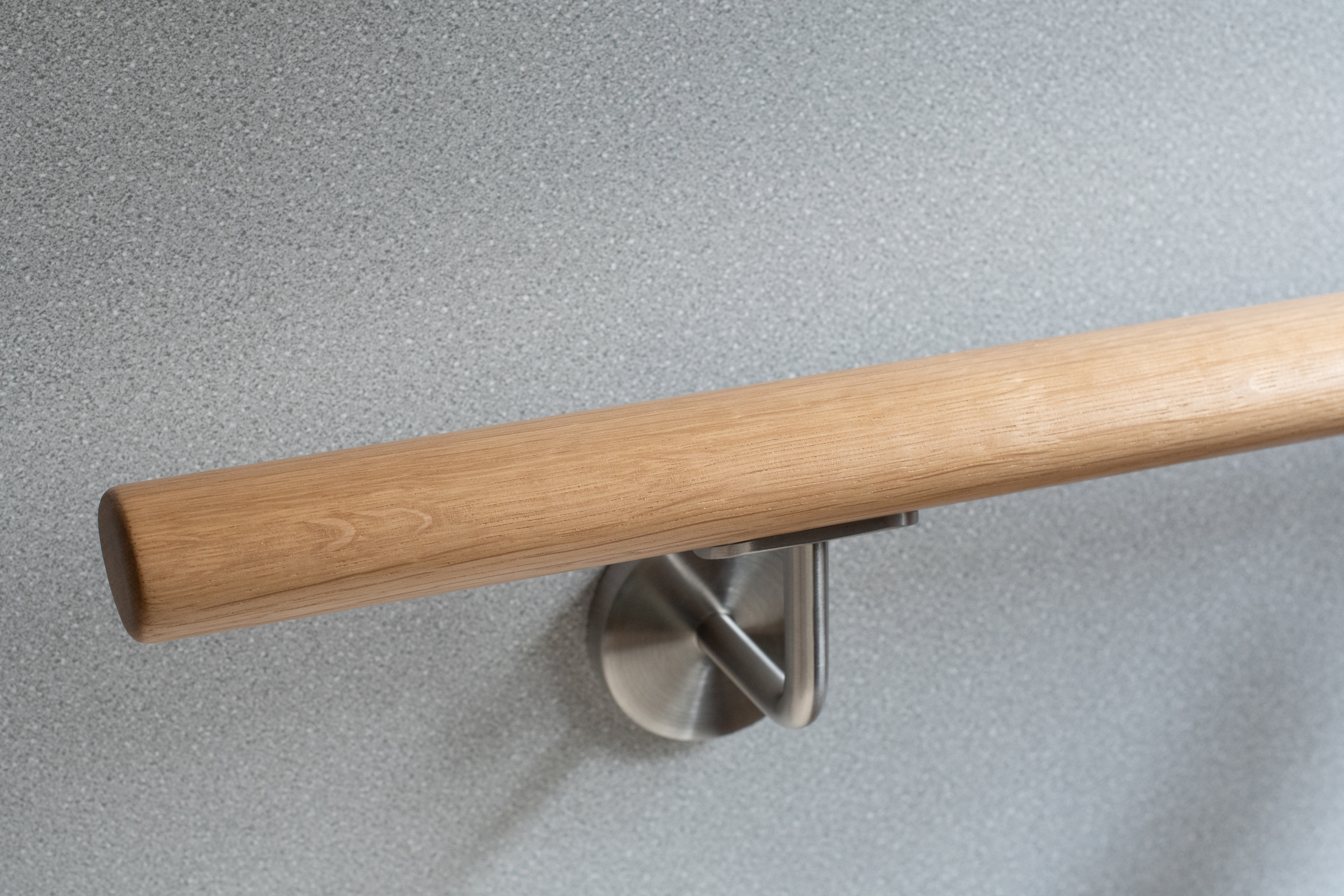 Pasamanos redondo de roble con soporte de acero inoxidable ante pared gris claro, primer plano con marcado veteado de roble