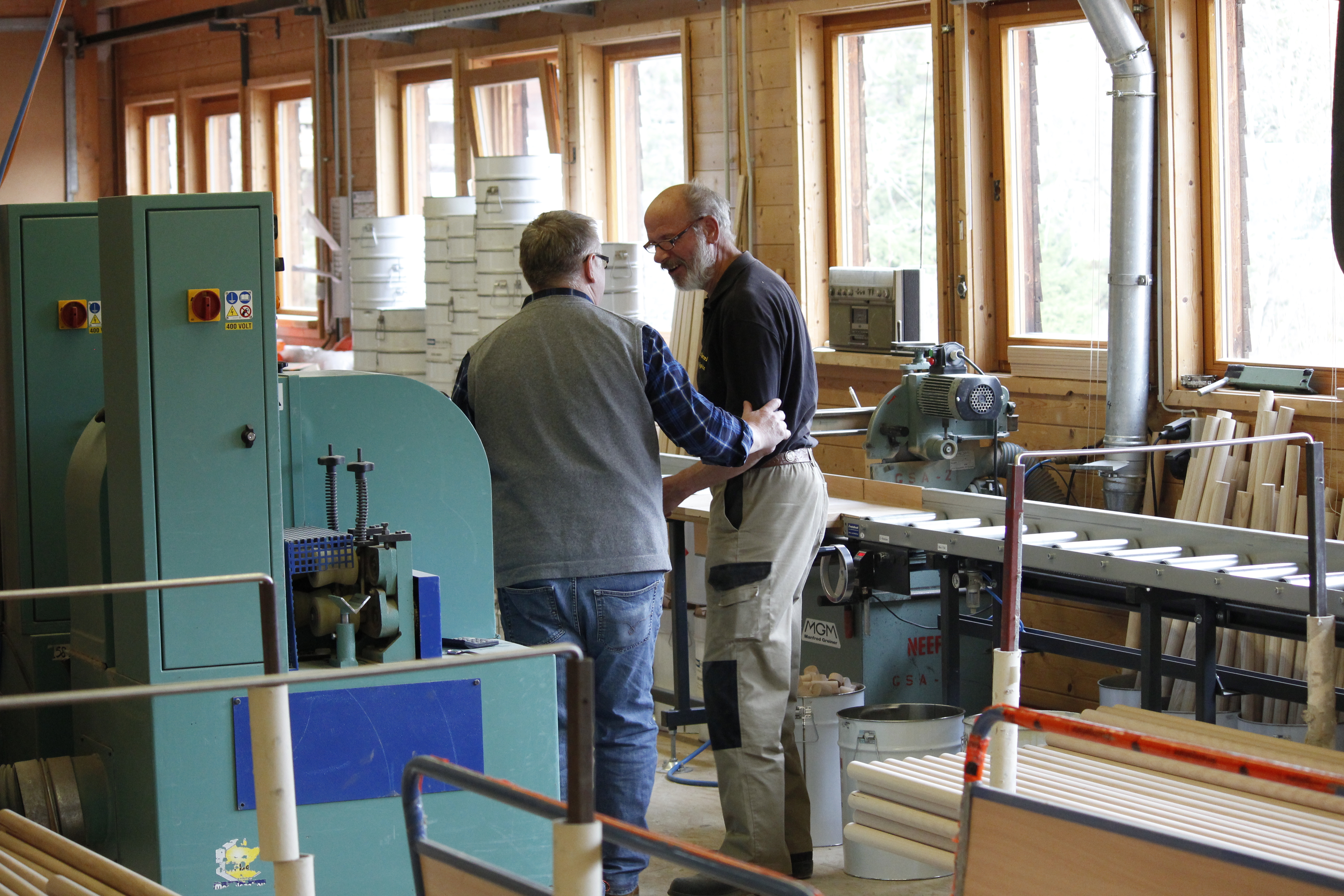 Dos empleados del taller de madera conversan entre máquinas, con ventanal y material de madera al fondo