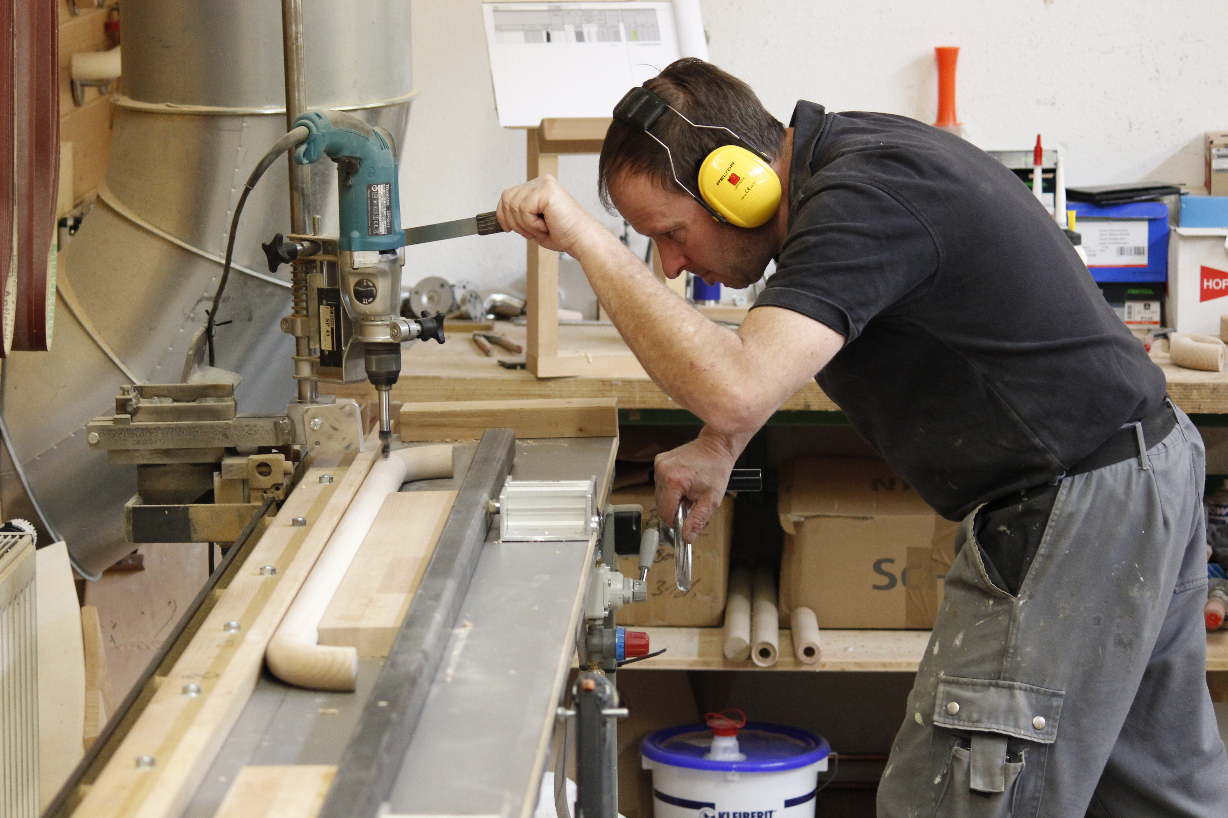 Artesano con protección auditiva taladra agujeros en un pasamanos de madera con una taladradora de columna en el taller