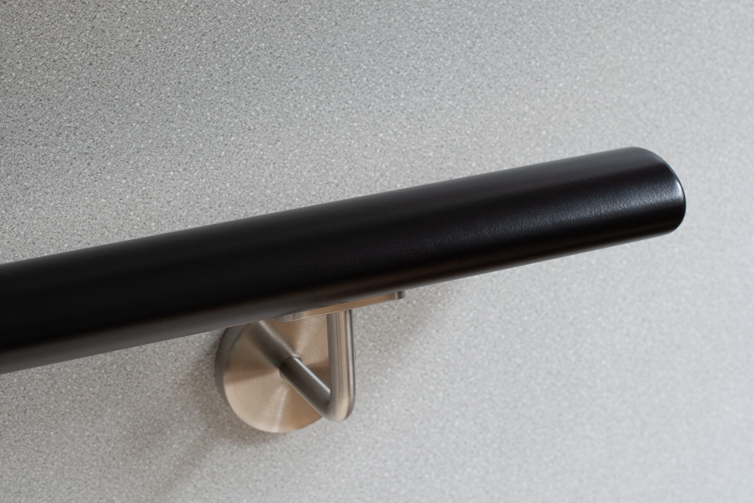 Pasamanos redondo de madera lacado en negro con soporte de acero inoxidable ante pared gris, detalle del extremo redondeado