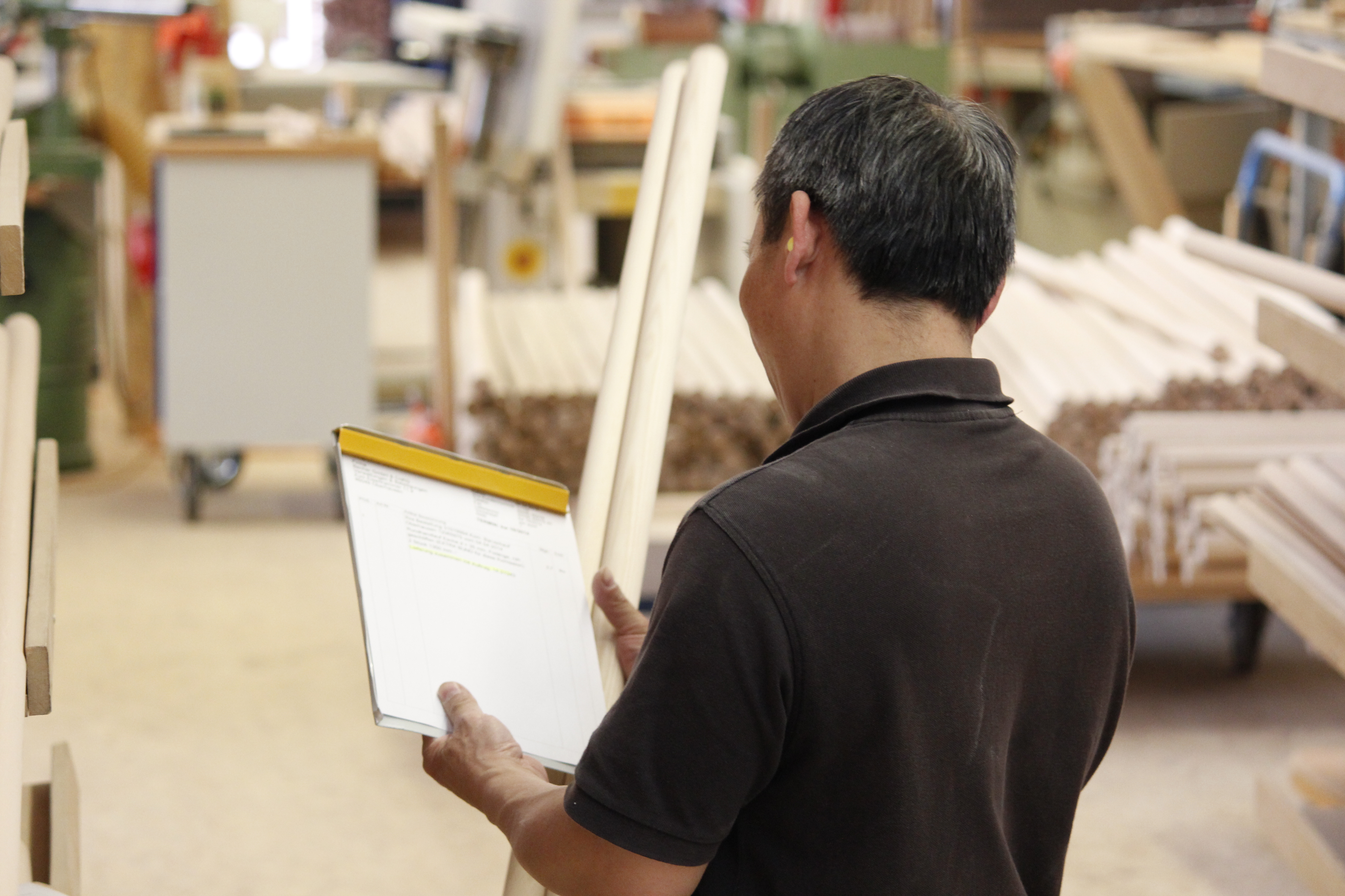 Empleado con carpeta de pedidos en el taller, barras redondas y piezas de pasamanos en bruto al fondo, vista de espaldas