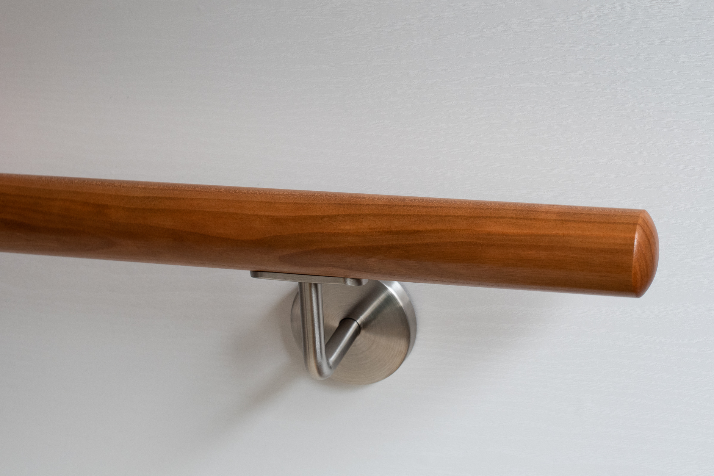 Pasamanos redondo de cerezo con soporte de acero inoxidable en pared blanca, madera marrón rojiza cálida, detalle del extremo