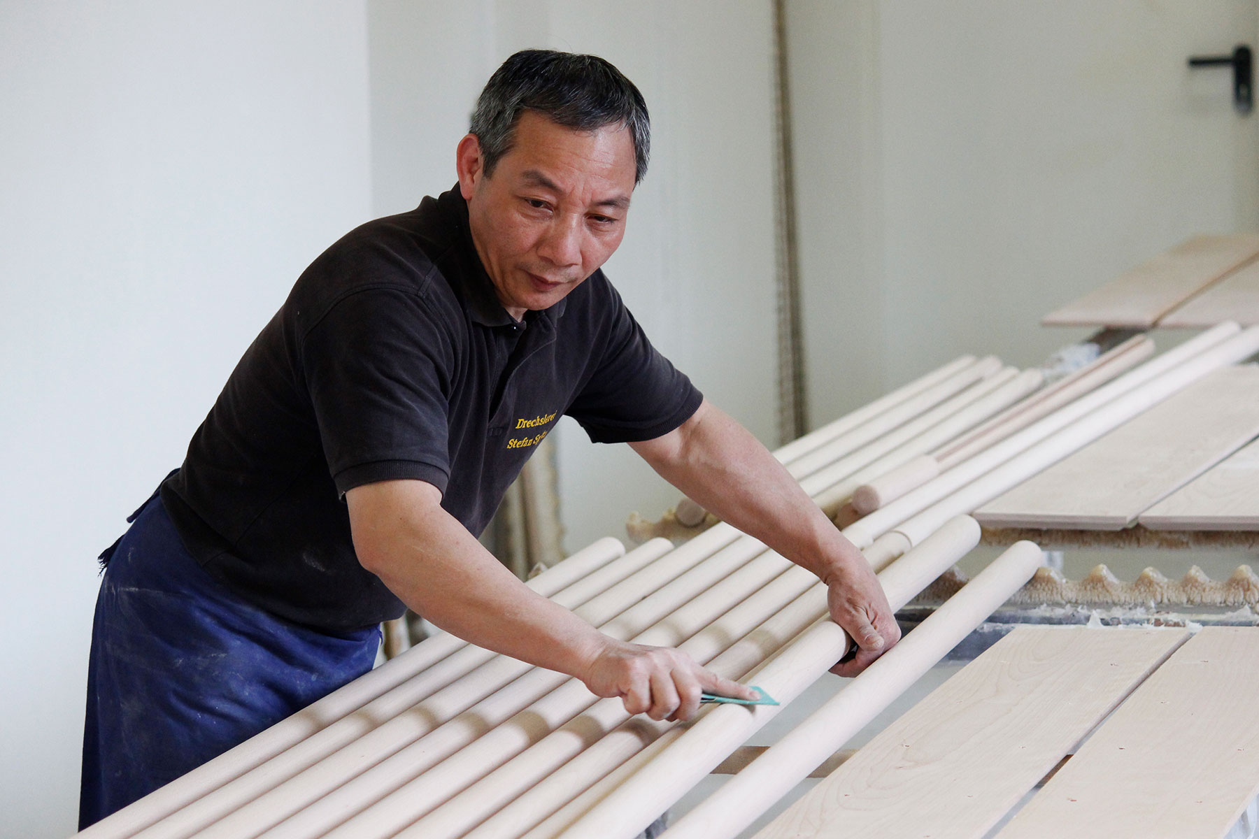 Fabricación de pasamanos: empleado revisa barras redondas de madera clara para pasamanos en el taller, tablones al fondo